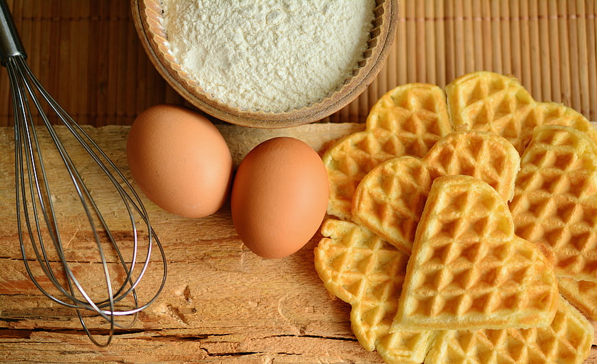 Heart-shaped waffles with eggs, flour, and whisk on wood