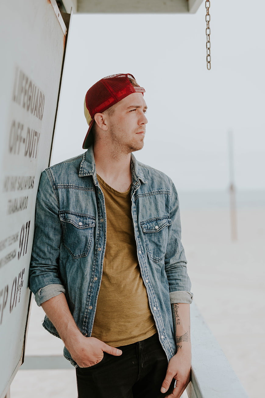 Man in Denim Jacket and Red Cap Leaning on Lifeguard Tower