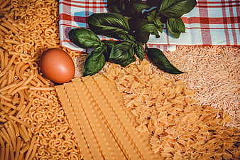 Flat lay of raw pasta shapes, brown egg, and fresh basil on checkered cloth
