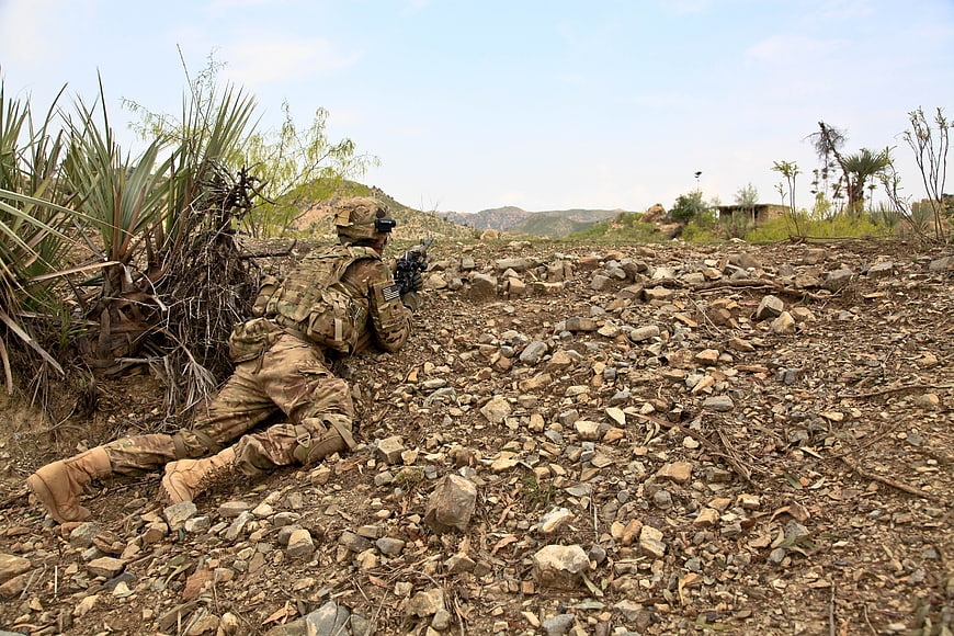 US Army Soldier In Combat Gear Crouching In Rocky Terrain