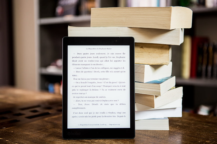Kobo e-reader displaying text next to a stack of hardcover books on a wooden table