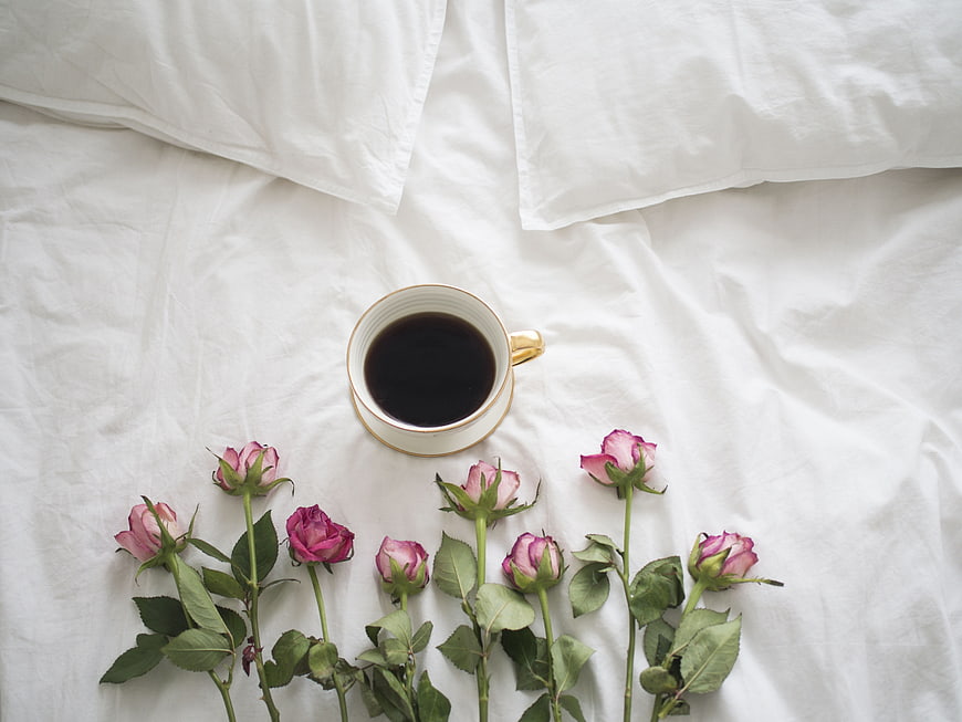 Flat lay of coffee cup and pink roses on white bed