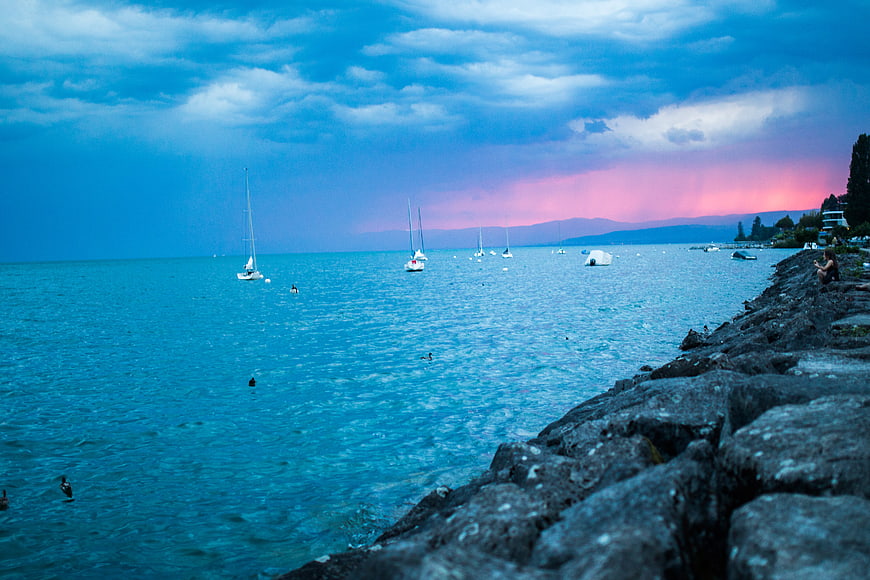 Dramatic Sunset Over Lake With Sailboats And Rocky Shore