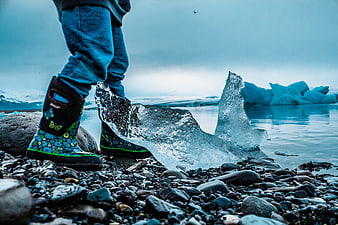 Child in Boots Standing by Blue Iceberg on Rocky Shore
