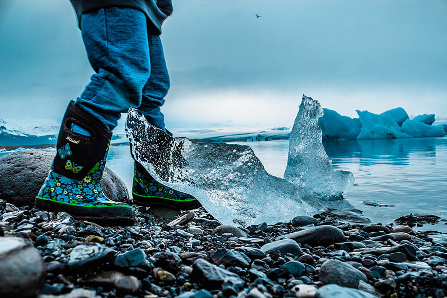 Child in Boots Standing by Blue Iceberg on Rocky Shore