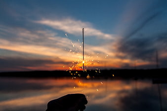 Hand holding sparkler against sunset lake background