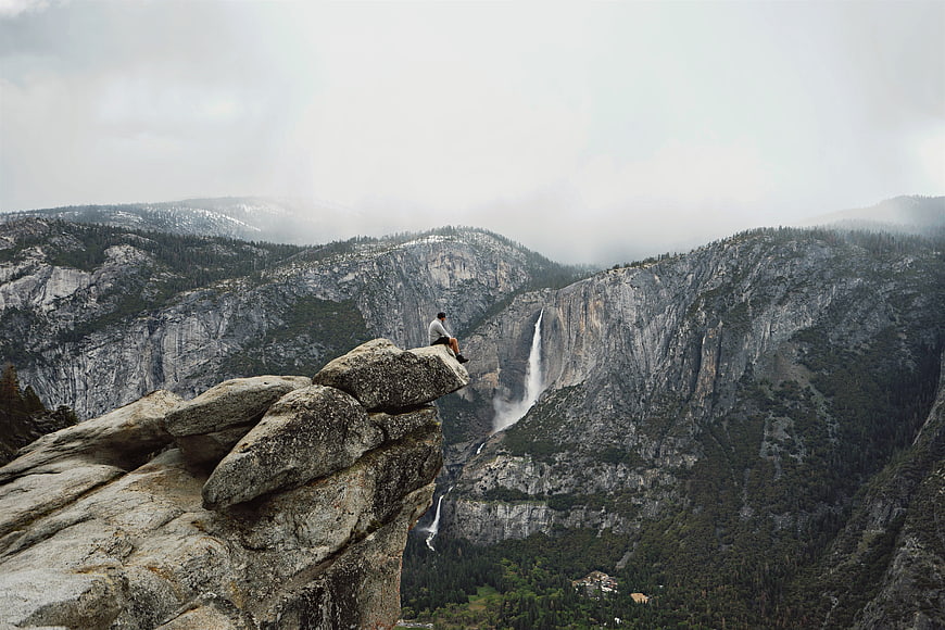 Man sitting on cliff edge overlooking waterfall and mountains