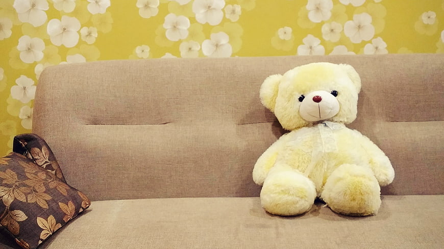 Yellow teddy bear sitting on beige sofa with floral wallpaper