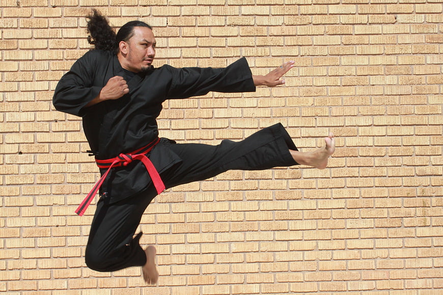 Male martial artist performing a dynamic jumping kick against a beige brick wall