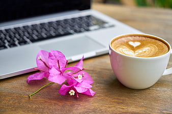 Latte with heart art and pink flowers beside laptop