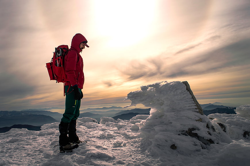Mountaineer in Red Jacket Standing on Snowy Mountain Peak at Sunset