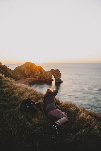 Man photographing coastal arch at sunrise from grassy cliff