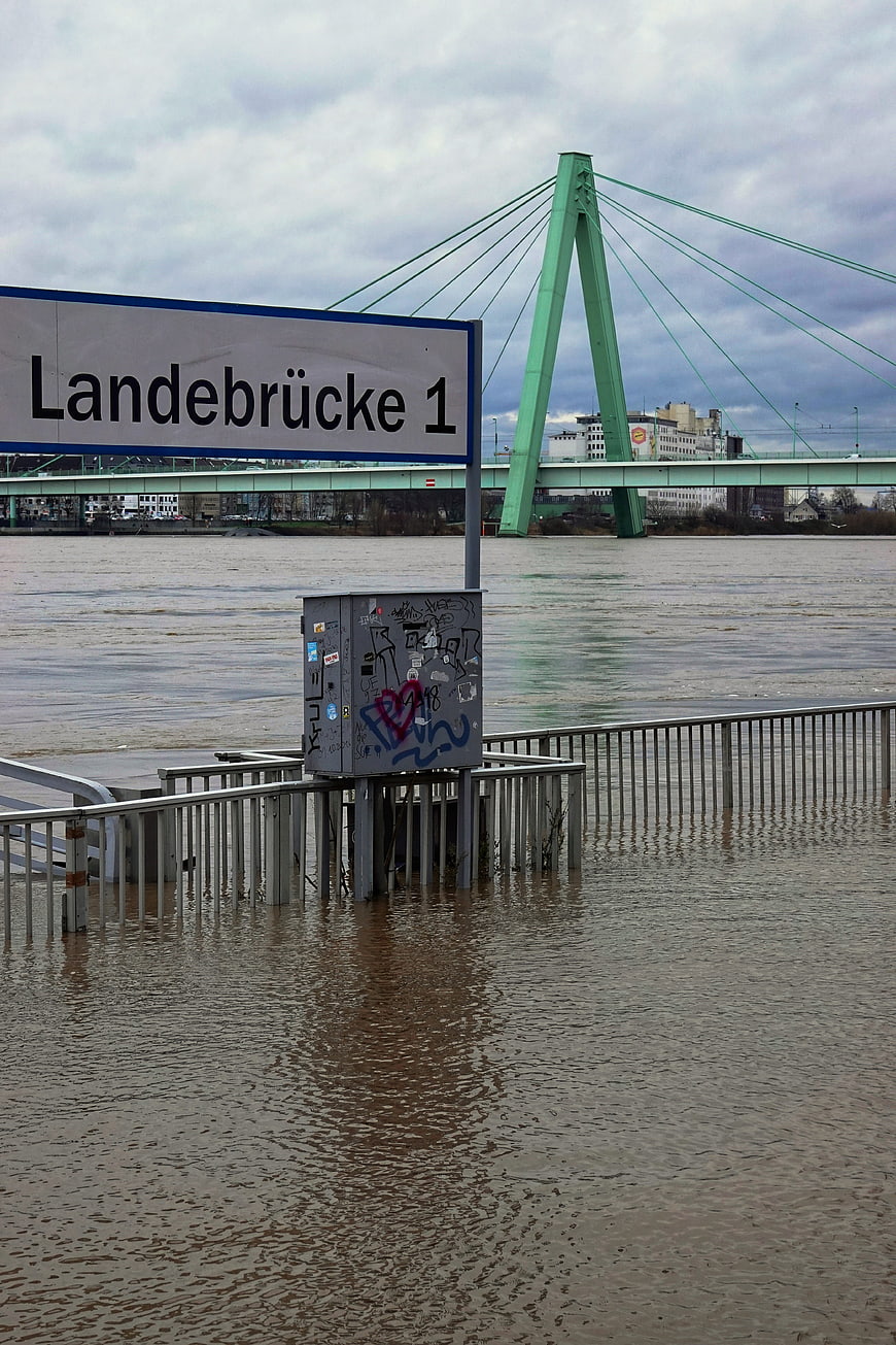 Flooded riverbank with Landebrücke 1 sign and green bridge