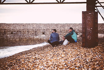 Two Men Relaxing on Pebble Beach Under Steel Bridge