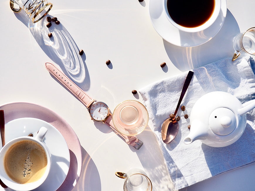 Flat lay of coffee, teapot, and pink watch on white table