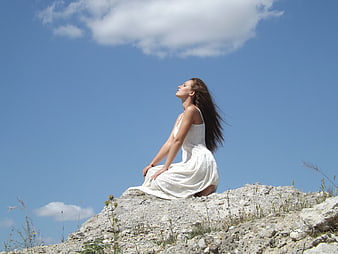 Woman in white dress kneeling on rocky hill under blue sky