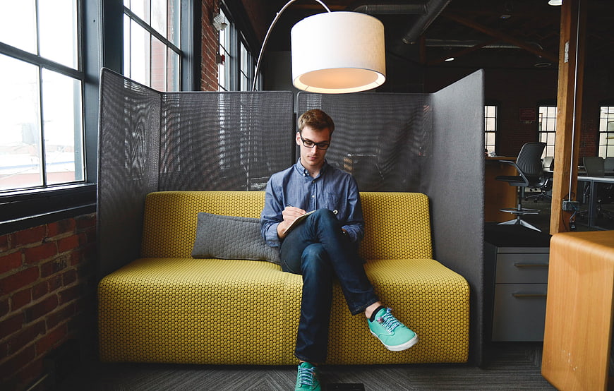 Young man writing in notebook on yellow sofa in modern office