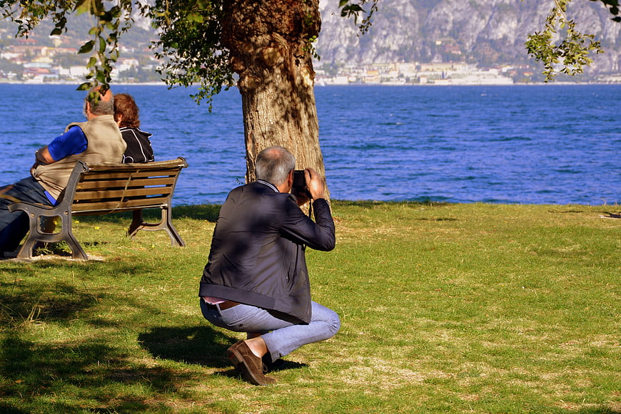 Senior man photographing couple on bench by lake