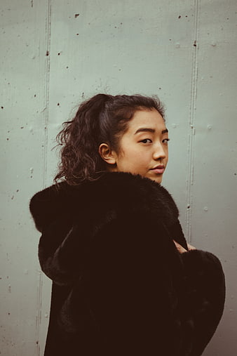 Asian woman in black fur coat looking back against concrete wall
