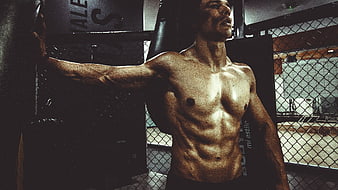 Muscular shirtless man posing in a dark gym with chain-link fence