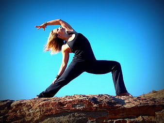 Woman doing yoga warrior pose on rocky cliff against blue sky
