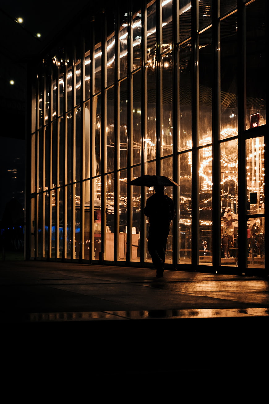 Silhouette of Person with Umbrella Walking Past Lit Glass Building at Night