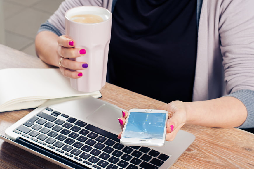 Woman holding coffee and smartphone at laptop workspace