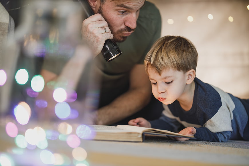 Father and son reading book with flashlight indoors
