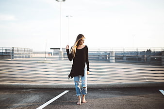 Barefoot woman in black top and ripped jeans walking on parking garage