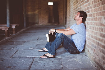 Man sitting against brick wall holding book in contemplation