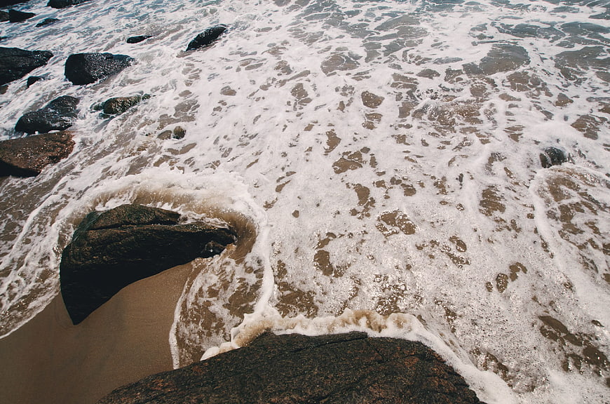 Foamy ocean waves crashing over dark rocks on sandy shore