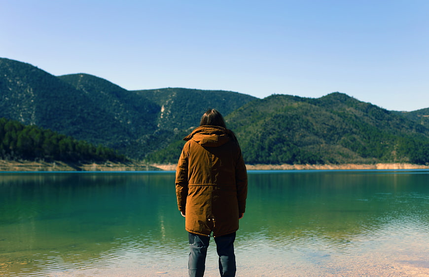 Man in Brown Coat Standing by Lake with Mountains