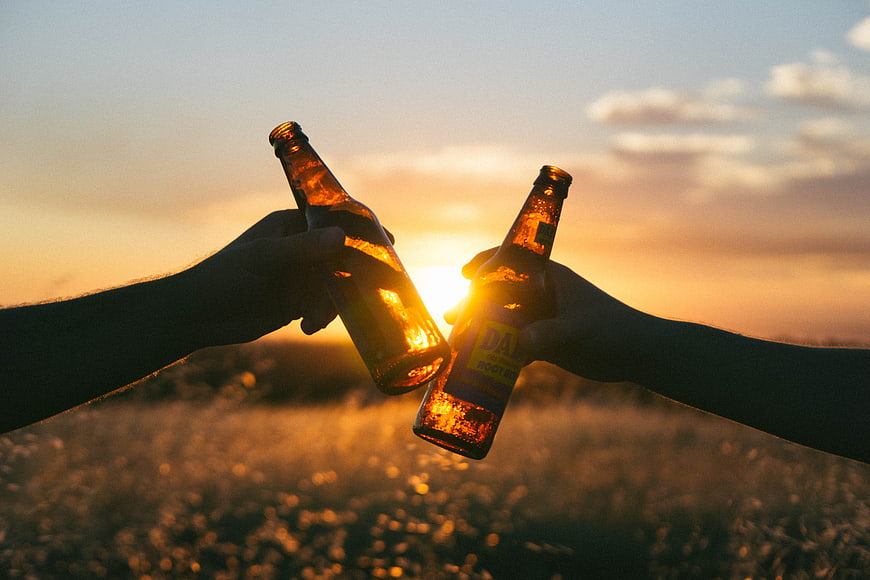 Two hands clinking beer bottles at sunset in a field