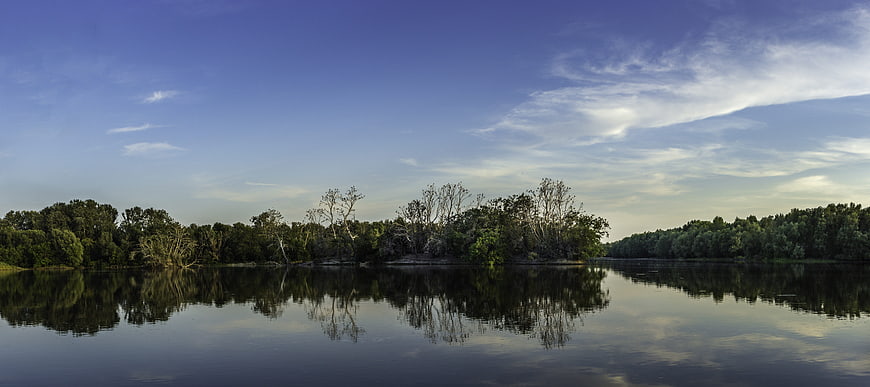 Panoramic view of calm river with tree reflections and blue sky