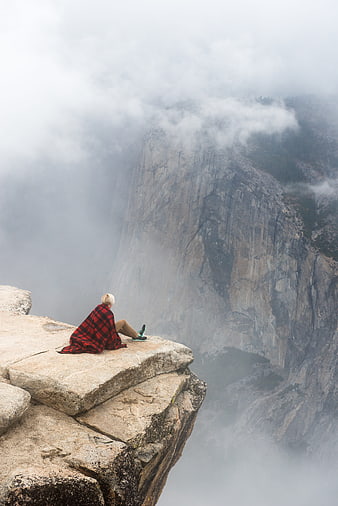 Man in red plaid blanket sitting on misty mountain cliff edge
