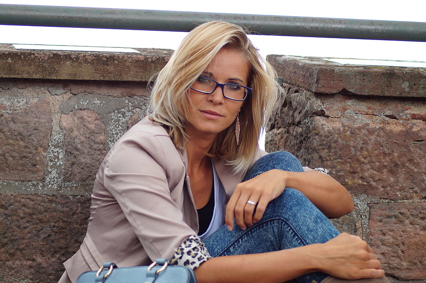 Blonde woman with glasses sitting against a rustic stone wall