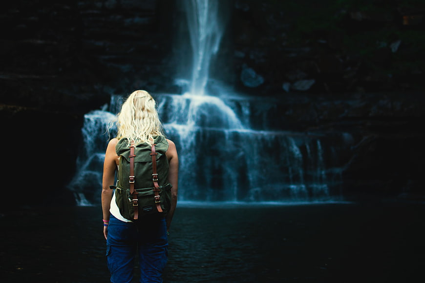 Woman with Backpack Standing Before Dark Waterfall