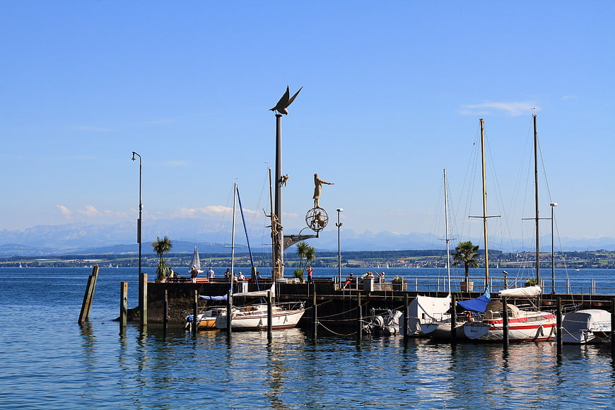 Sailboats docked at Meersburg harbor with bird sculpture and mountains