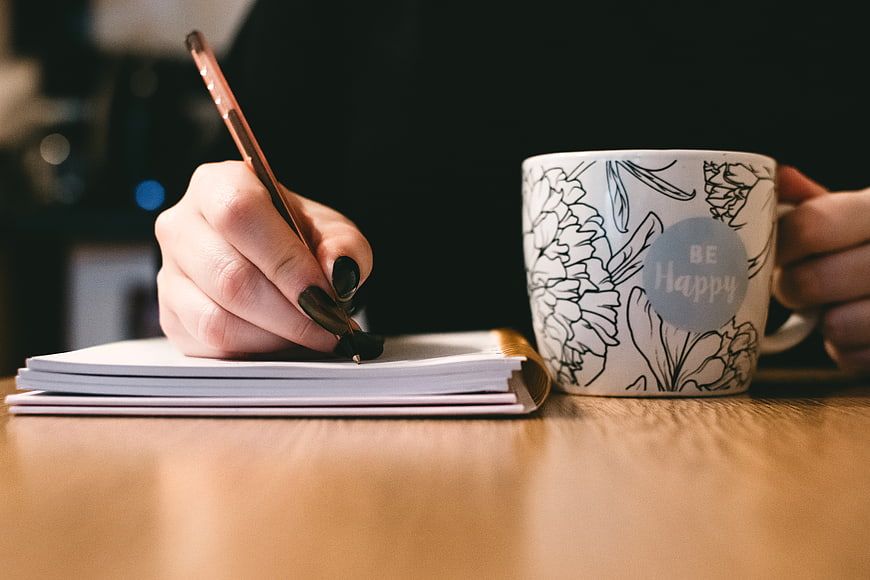 Woman Writing in Notebook with Be Happy Coffee Mug