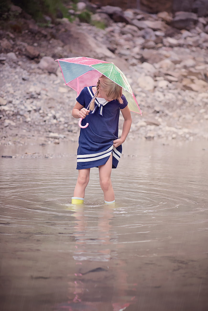 Young Girl with Rainbow Umbrella Standing in Rainy Stream