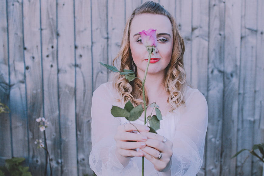 Blonde Woman Holding Pink Rose Against Wooden Fence