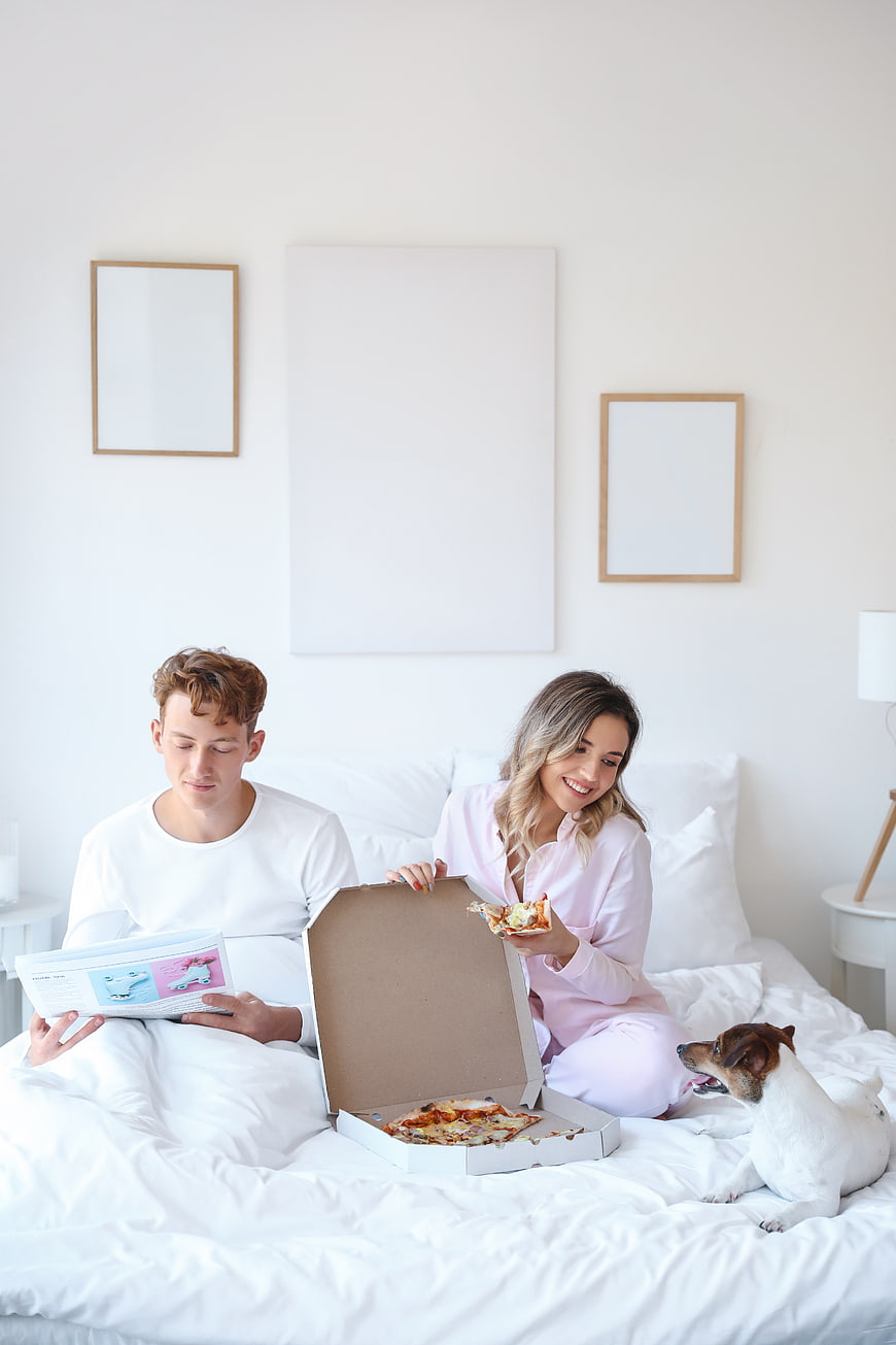 Happy couple eating pizza in bed with dog
