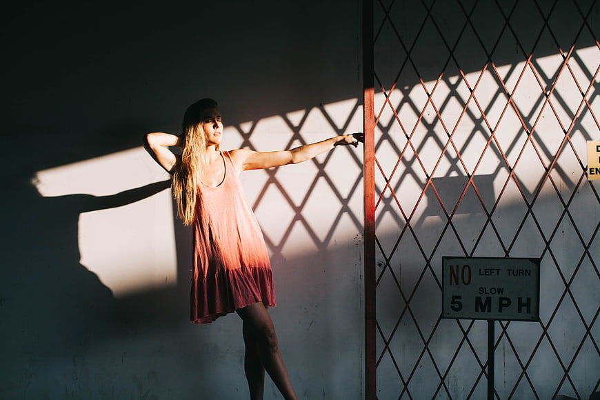 Woman in Pink Dress Leaning Against Wall with Fence Shadows