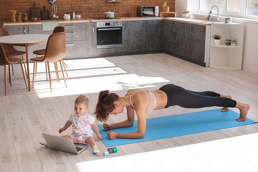 Mother doing yoga plank while toddler watches laptop in kitchen