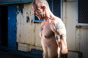 Shirtless muscular man with tattoos grimacing during intense outdoor workout