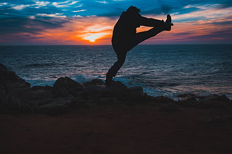 Silhouette of person stretching on rocky coast at sunset
