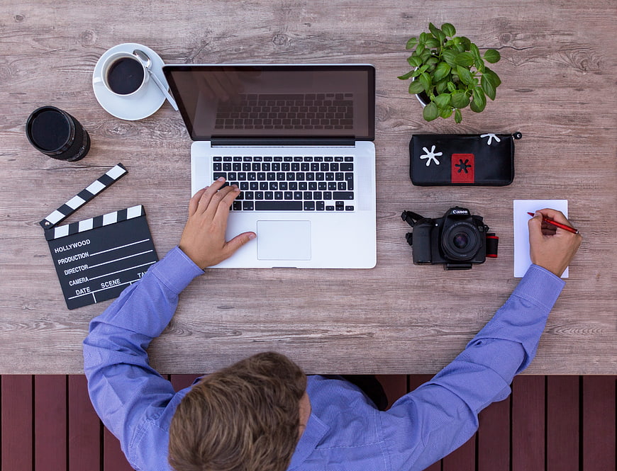 Top view of filmmaker working at wooden desk with laptop and camera
