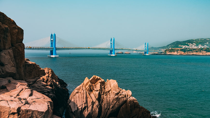 Blue suspension bridge over ocean with rocky foreground