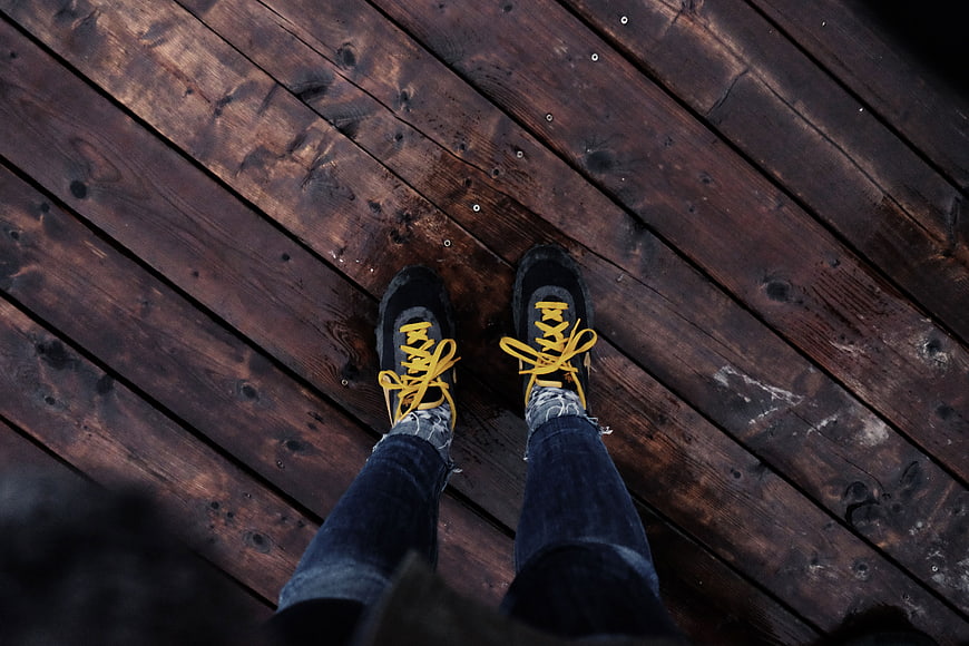 First-person view of black sneakers with yellow laces on wooden deck