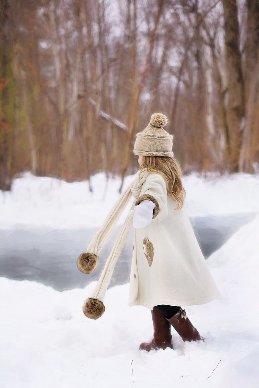 Little girl in cream winter coat and hat walking in snowy forest
